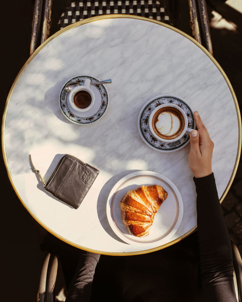 Italienischer Frühstücksmoment auf einer Marmortischplatte mit Croissant, Cappuccino mit Latte Art und klassischem Espresso in stilvollen Porzellantassen.