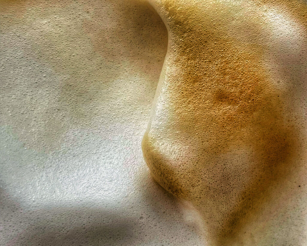 Makroaufnahme von feinporigem Milchschaum eines frisch zubereiteten Cappuccinos, der die cremige Konsistenz und goldbraune Farbe des Schaums zeigt.