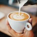 Detailaufnahme eines Cappuccinos mit Latte Art, während frische Milch aus einem Kännchen in die Tasse gegossen wird – ein Moment der Barista-Kunst.