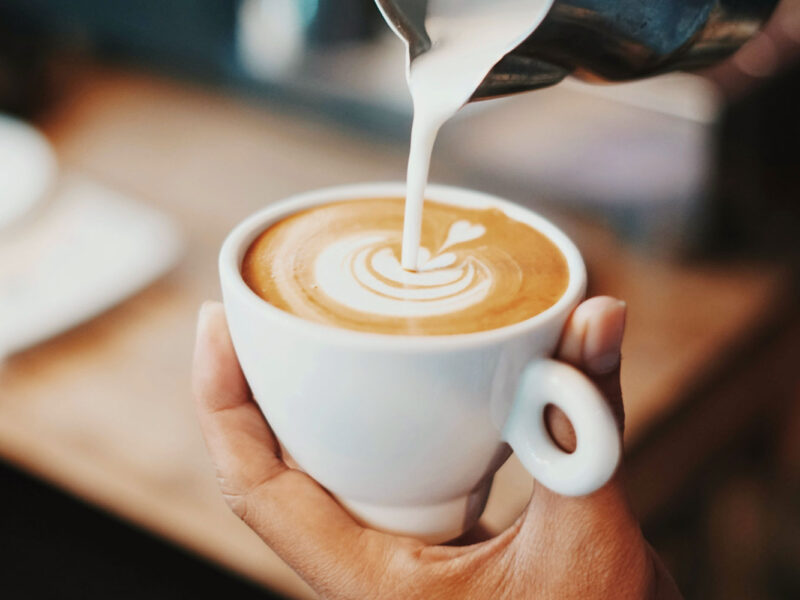 Detailaufnahme eines Cappuccinos mit Latte Art, während frische Milch aus einem Kännchen in die Tasse gegossen wird – ein Moment der Barista-Kunst.