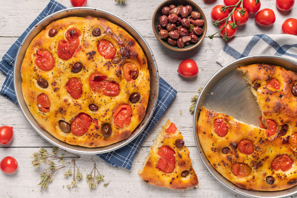 Zwei runde Bleche mit traditioneller Focaccia Barese, belegt mit Tomaten und Oliven, daneben frische Zutaten auf weißem Holztisch.