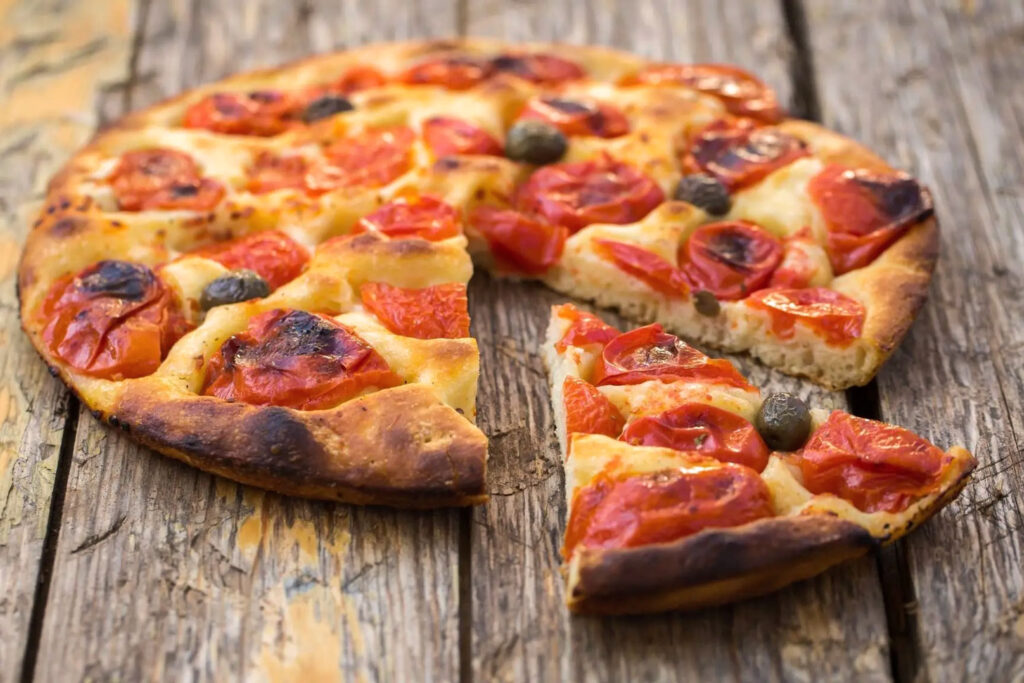 Focaccia Barese mit kräftig gebackener Kruste, roten Tomaten und Kapern auf rustikalem Holztisch serviert.