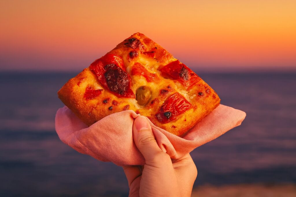 Ein Stück Focaccia mit Paprika, Tomaten und grüner Olive, im Sonnenuntergang am Meer genossen – italienisches Street Food pur.