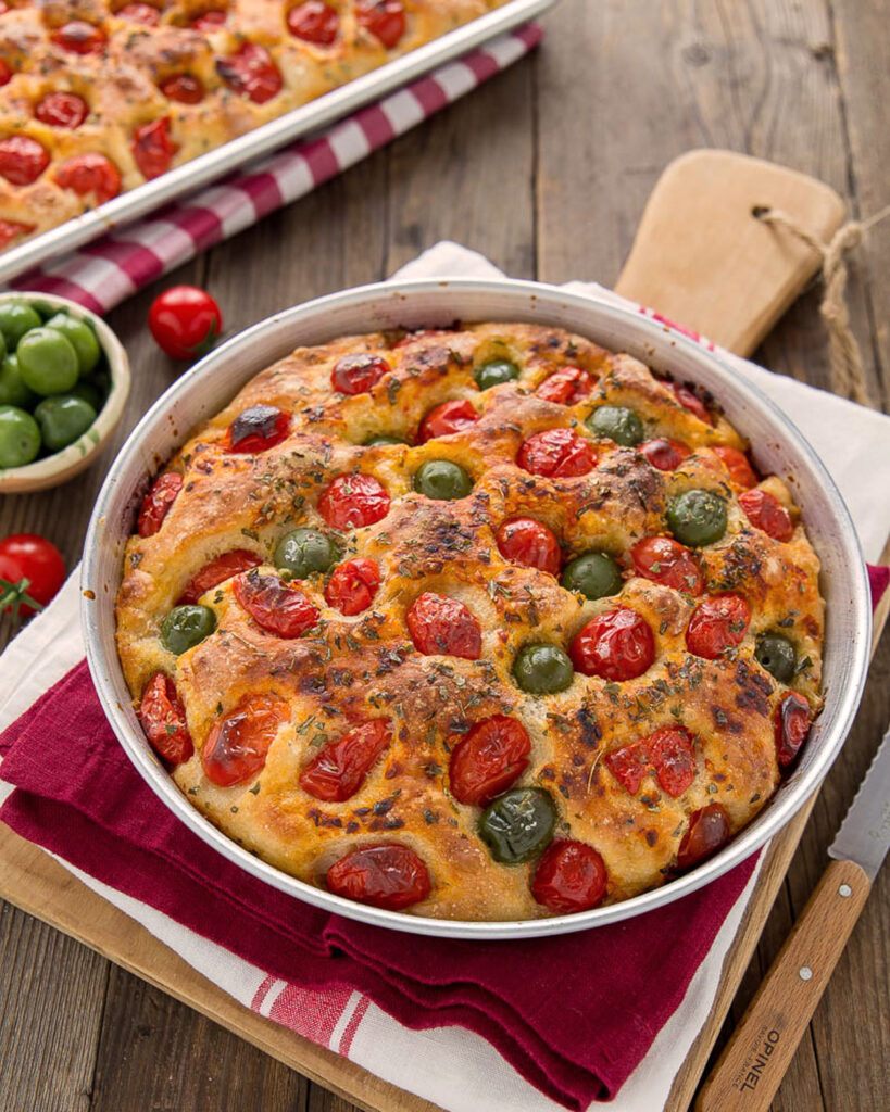 Focaccia Barese mit Kirschtomaten und grünen Oliven in runder Backform auf rustikalem Holztisch.