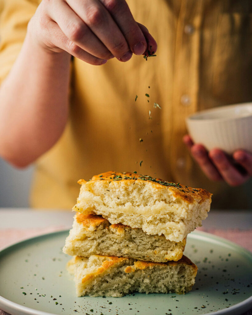 Hand bestreut frische Focaccia-Stücke mit Kräutern, angerichtet auf einem hellen Teller.