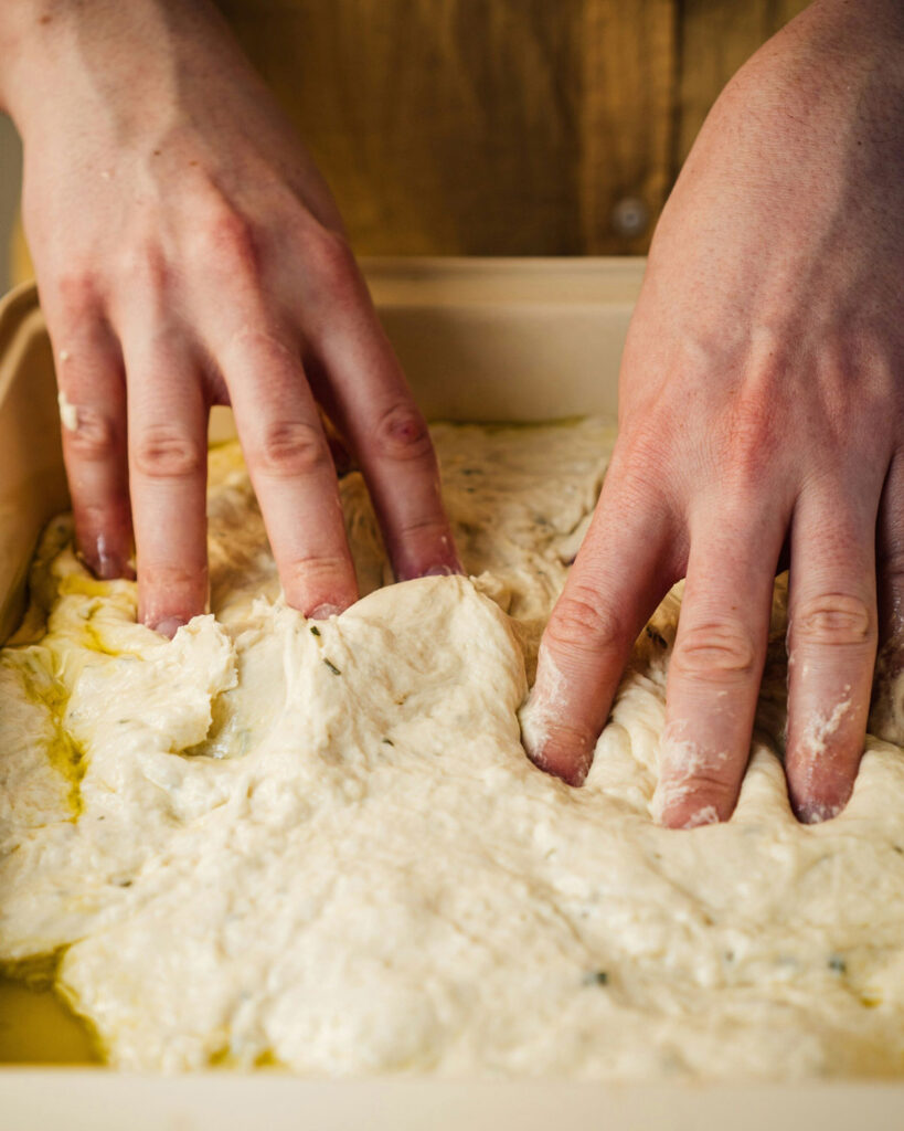 Hände drücken den Focaccia-Teig in eine Form, mit Olivenöl bedeckt.