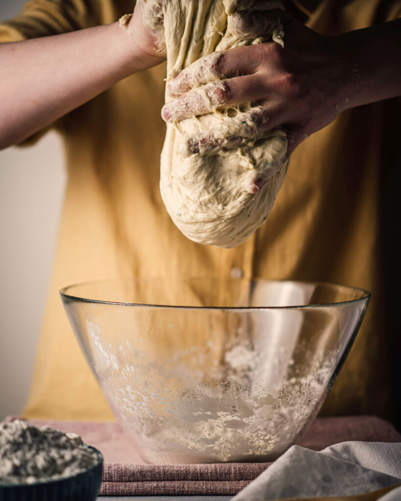 Focaccia-Teig wird mit bemehlten Händen aus einer Glasschüssel herausgehoben.