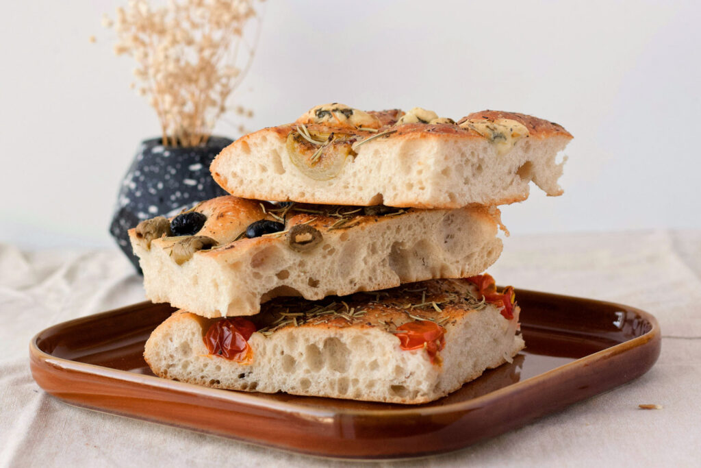 Drei rustikale Stücke Focaccia mit Oliven, Tomaten und Rosmarin, gestapelt auf einem Teller.
