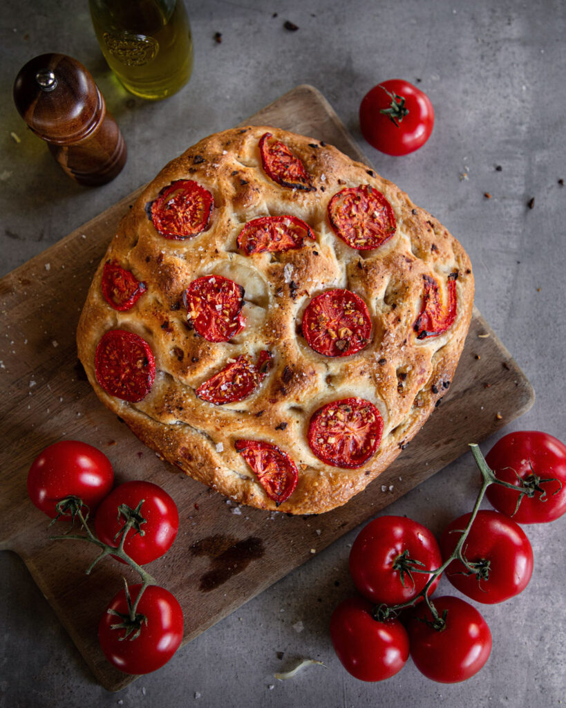 Runde Focaccia mit frischen Tomatenscheiben auf einem Holzbrett, umgeben von Strauchtomaten.