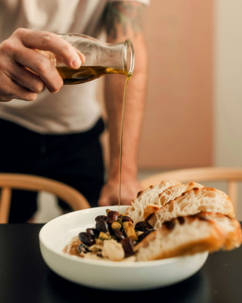 Hand gießt hochwertiges Olivenöl über eine Schale mit Brot, Bohnen und eingelegtem Gemüse.