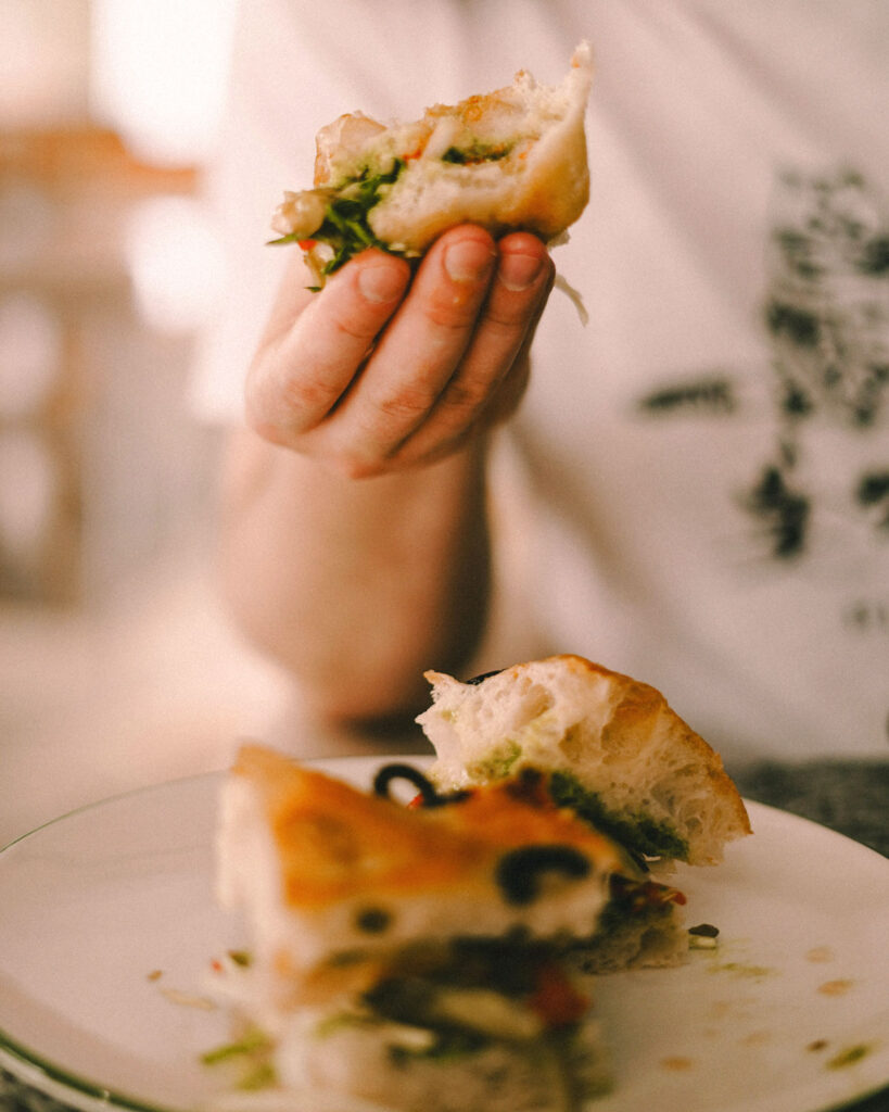 Person hält ein Stück Focaccia-Sandwich mit Oliven und frischem Grün in der Hand.