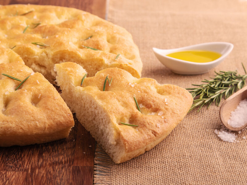Runde Focaccia mit Rosmarin, grobem Salz und Olivenöl auf Holztisch, daneben frische Kräuter
