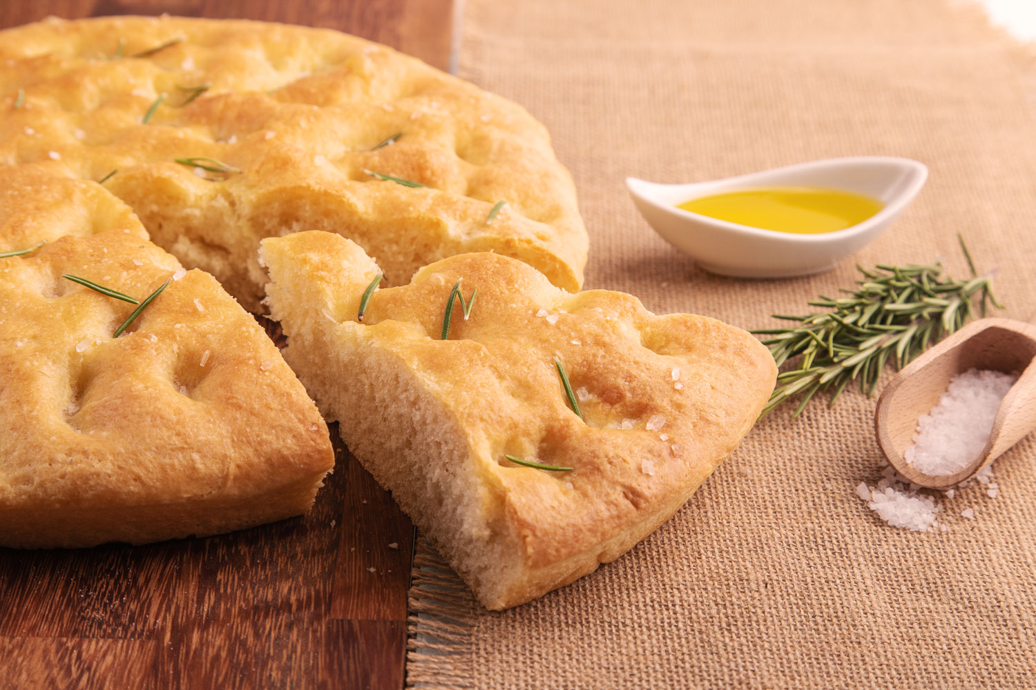 Runde Focaccia mit Rosmarin, grobem Salz und Olivenöl auf Holztisch, daneben frische Kräuter