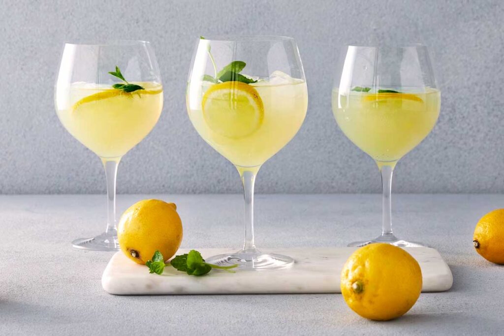 Drei Gläser Limoncello Spritz mit Zitrone und Minze serviert auf einem hellen Untergrund, daneben frische Zitronen.