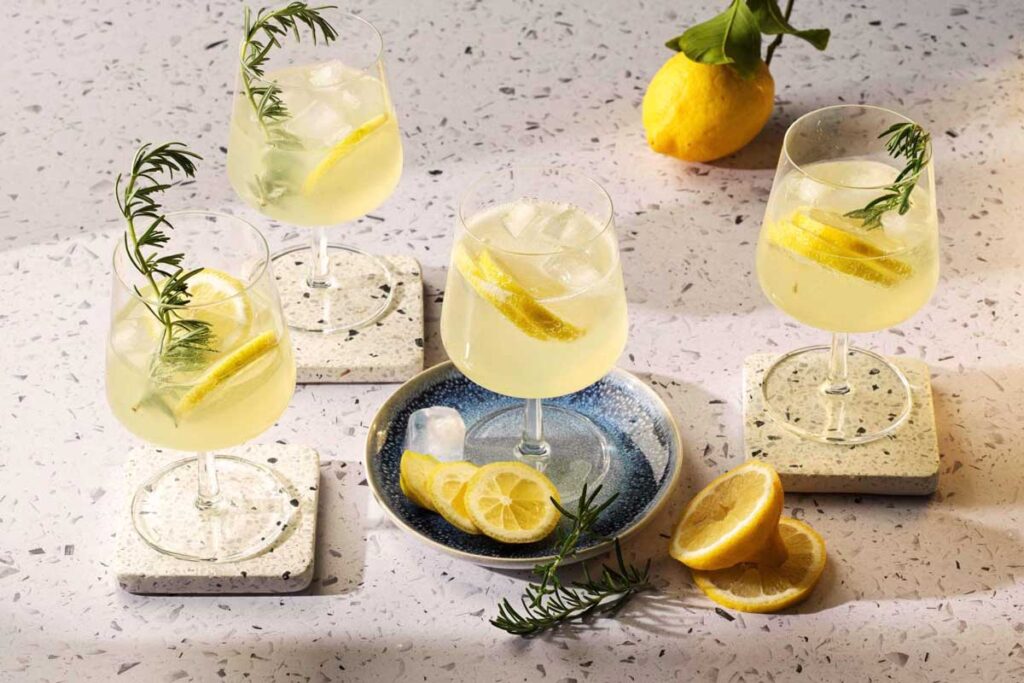 Vier Gläser erfrischender Limoncello Spritz auf dekorativen Untersetzern, garniert mit Zitrone und Rosmarin, daneben frische Zitronenscheiben.