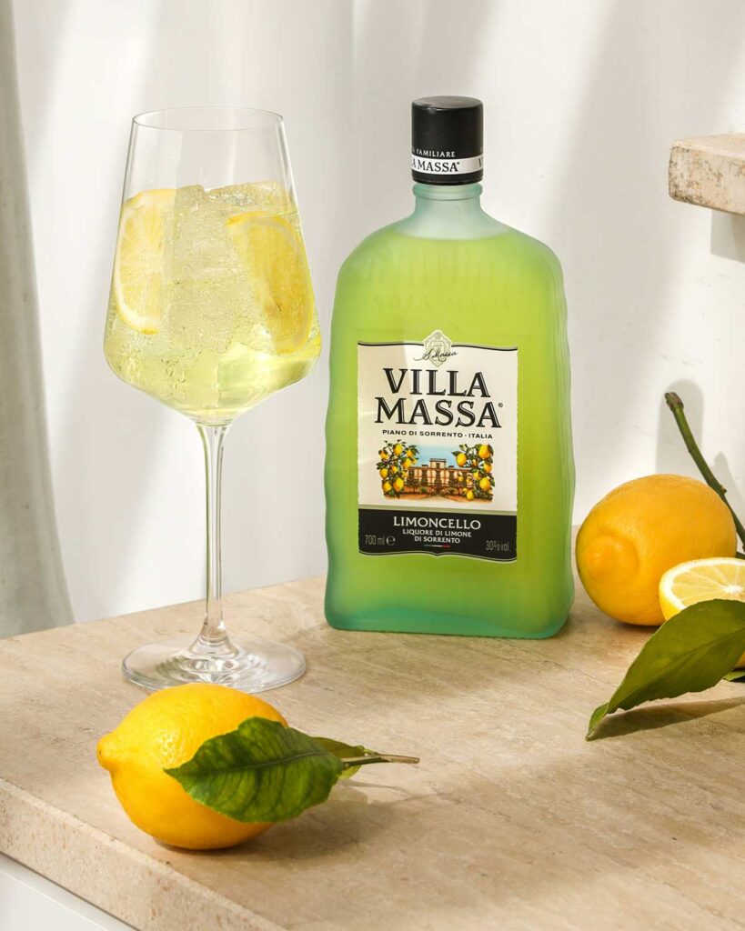 Flasche Villa Massa Limoncello neben einem Glas mit Limoncello Spritz, Eiswürfeln und Zitronenscheiben, dekoriert mit frischen Zitronen.