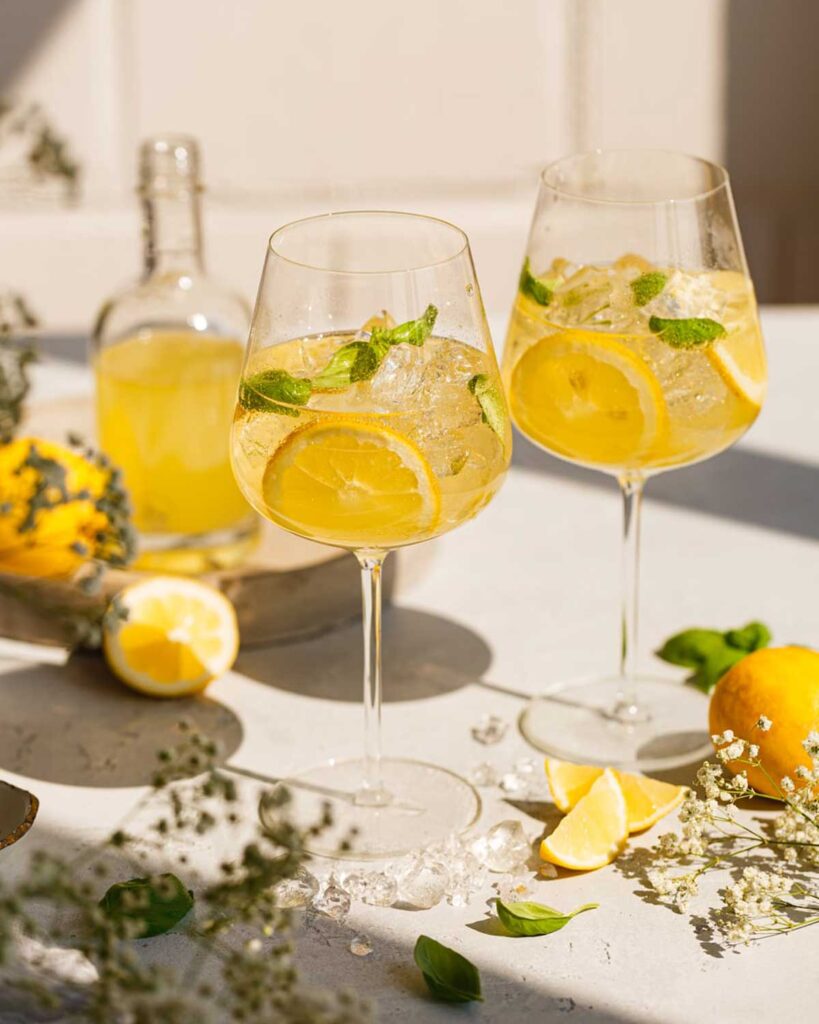 Zwei Gläser Limoncello Spritz auf einem sommerlich gedeckten Tisch, umgeben von Zitronen, Eiswürfeln, Basilikum und Blüten.