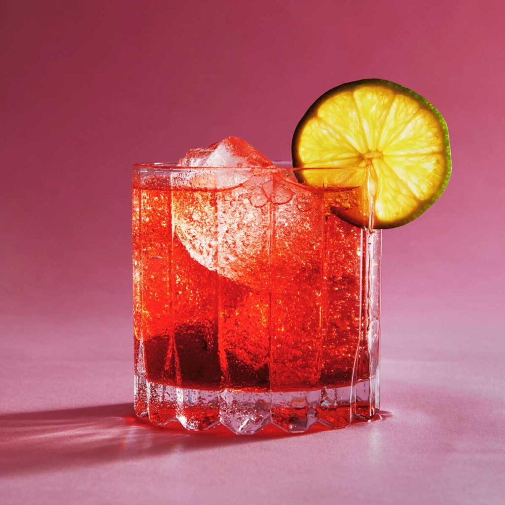 Leuchtend roter Negroni-Cocktail mit großer Eiskugel und Limettenscheibe am Glasrand, vor pinkem Hintergrund.