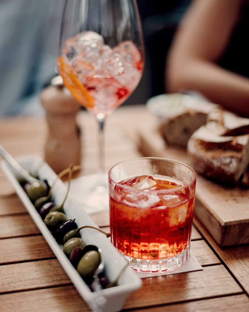 Negroni-Cocktail im Tumbler auf einem Holztisch, serviert zu Oliven und Aperitivo, im Hintergrund ein weiteres Glas mit Eis und Orangenzeste.