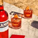 Zwei Gläser Negroni mit Eiswürfeln und Orangenscheiben auf einem Marmortisch am Strand, neben einer Flasche Campari.