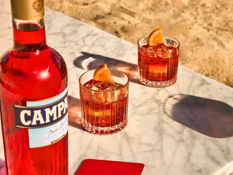 Zwei Gläser Negroni mit Eiswürfeln und Orangenscheiben auf einem Marmortisch am Strand, neben einer Flasche Campari.