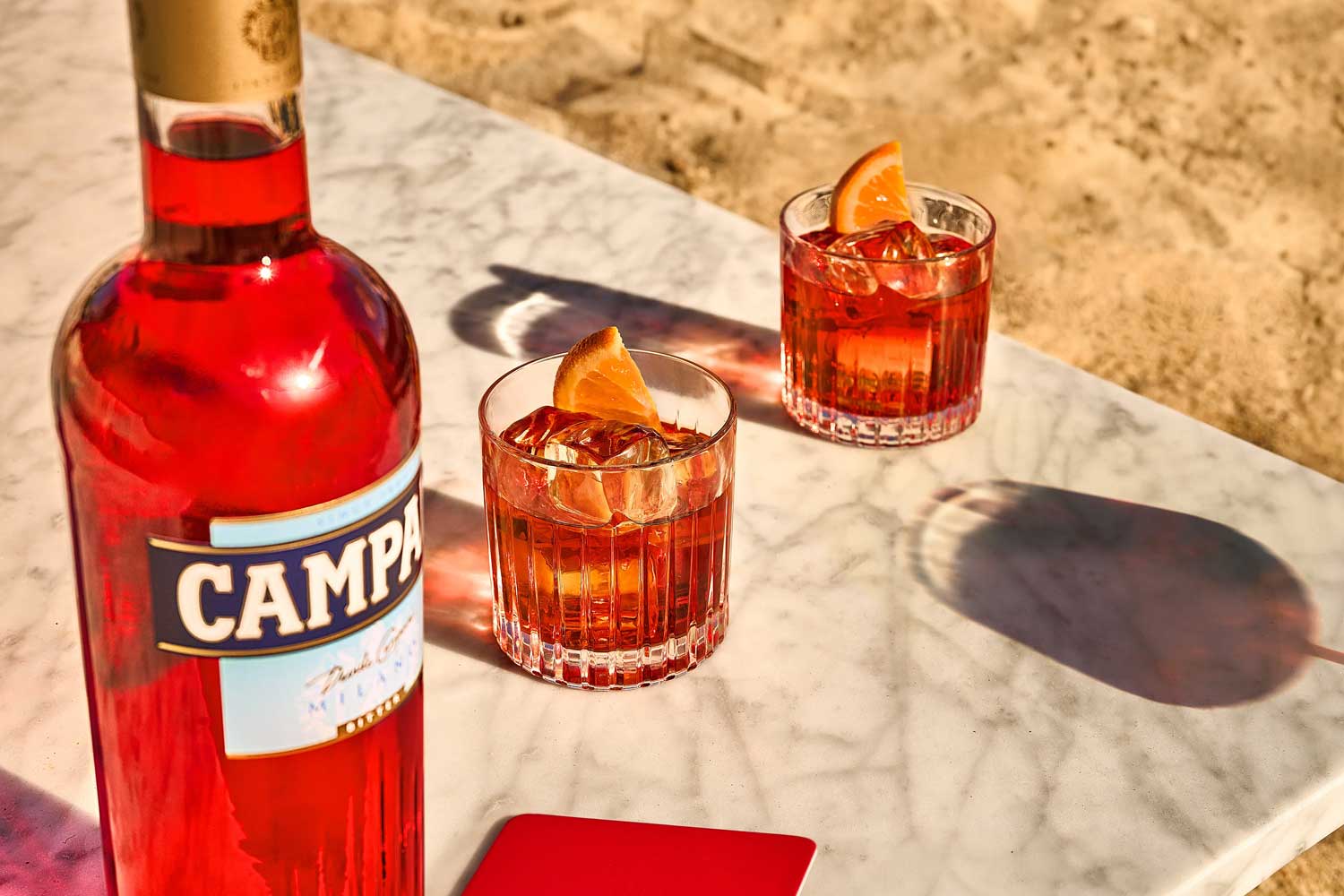 Zwei Gläser Negroni mit Eiswürfeln und Orangenscheiben auf einem Marmortisch am Strand, neben einer Flasche Campari.