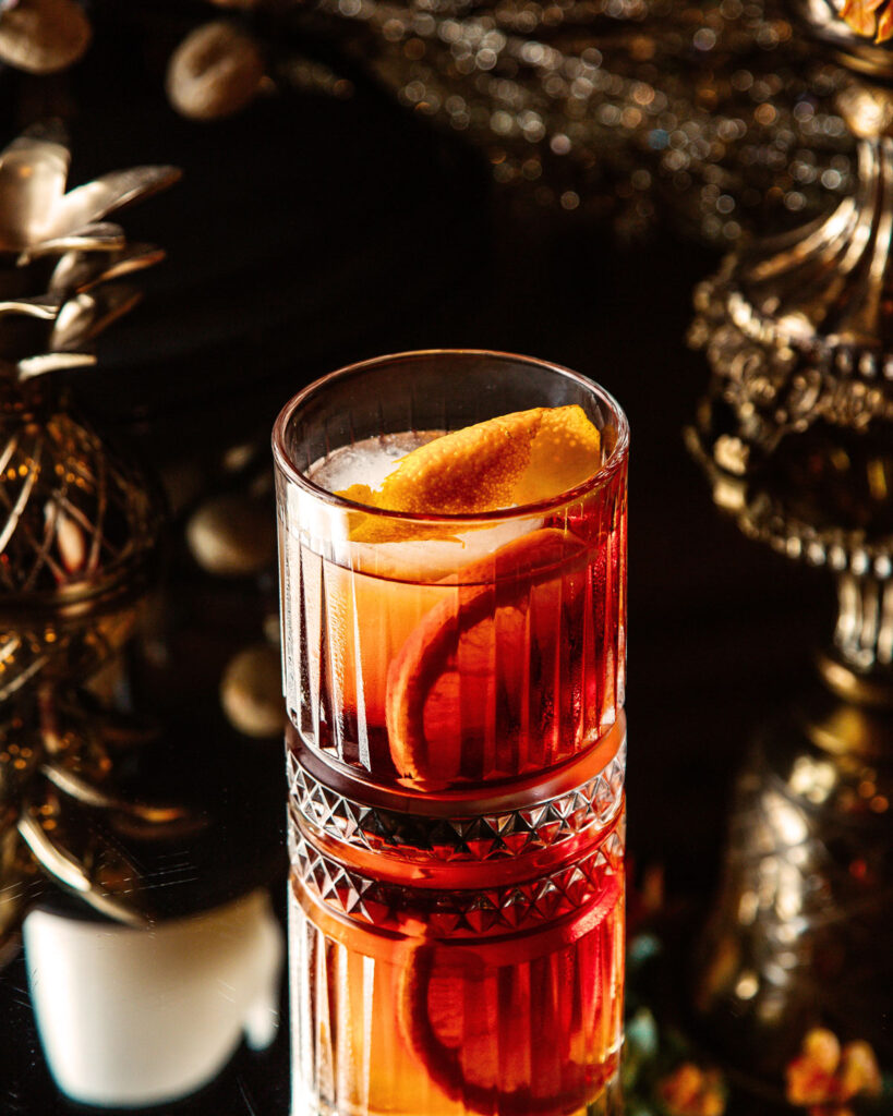 Negroni-Cocktail in einem geschliffenen Glas mit Eiswürfeln und Orangenzeste – serviert auf glänzendem Tisch mit eleganter, dunkler Dekoration.