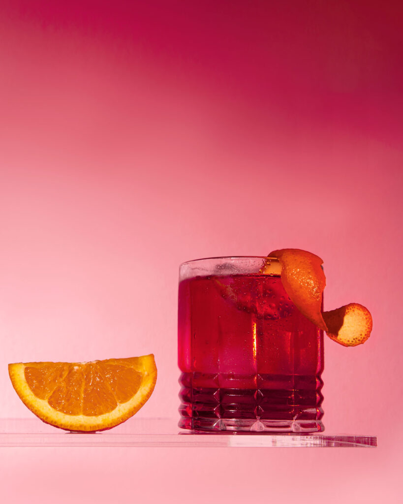 Negroni Sbagliato in schlichtem Glas mit Eiswürfeln, Orangenzeste und einer Orangenscheibe daneben – vor knallig pinkem Hintergrund.