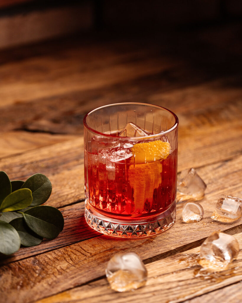 Negroni mit Orangenzeste und Eiswürfeln auf einem rustikalen Holztisch, umgeben von geschmolzenem Eis und grünem Blattwerk.