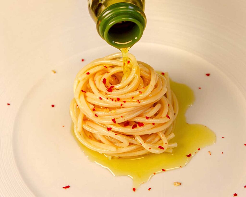 Spaghetti Aglio, Olio e Peperoncino mit Olivenöl direkt aus der Flasche verfeinert, minimalistisch angerichtet auf weißem Teller.