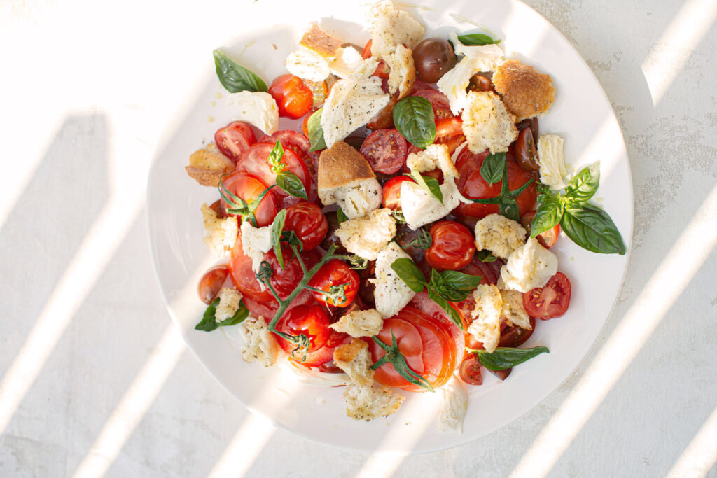 Neu interpretierter Caprese-Salat mit Tomaten, Mozzarella, Brotstückchen und Basilikum, im Sonnenlicht serviert.