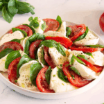 Modern angerichteter Caprese-Salat mit Tomaten, Mozzarella, frischem Basilikum und italienischen Kräutern auf weißem Teller.