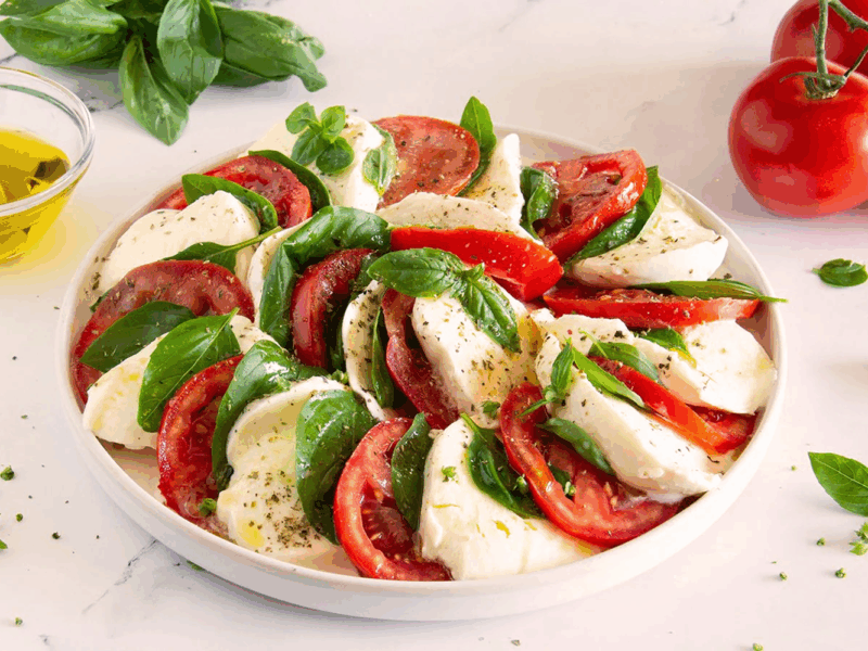 Modern angerichteter Caprese-Salat mit Tomaten, Mozzarella, frischem Basilikum und italienischen Kräutern auf weißem Teller.