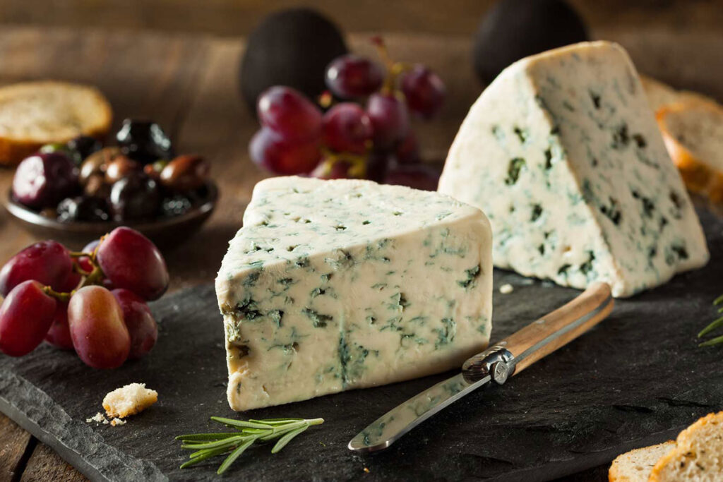 Zwei Stücke Gorgonzola auf einer Schieferplatte, angerichtet mit Trauben, Oliven und Rosmarin – perfektes Käsearrangement.
