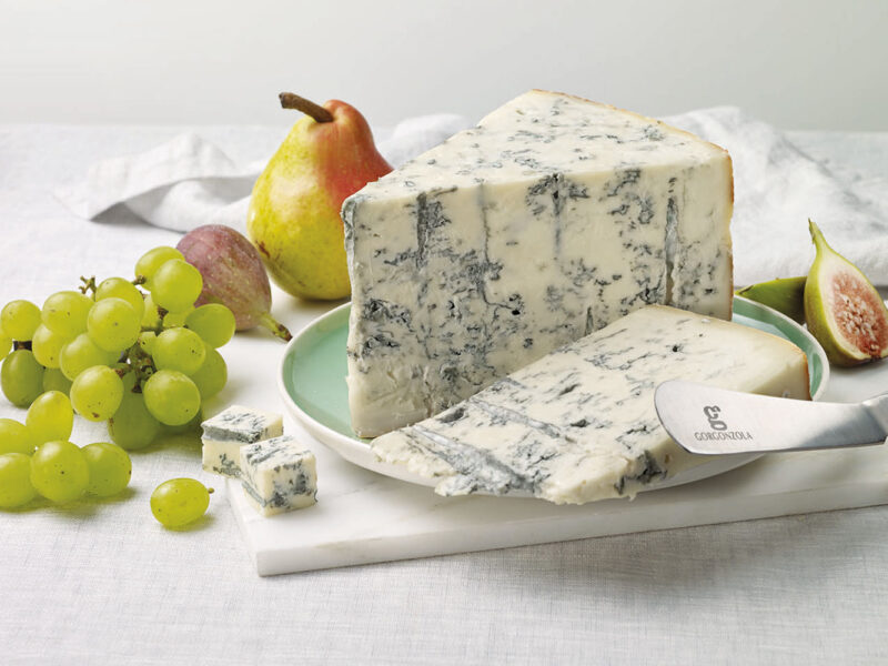 Großes Stück Gorgonzola auf einer hellen Platte, angerichtet mit frischen Trauben, Birne, Feigen und speziellem Käsemesser – Gorgonzola trifft Frische.