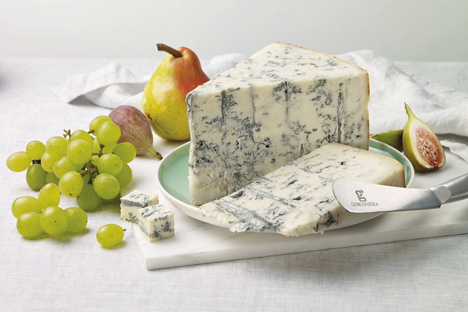 Großes Stück Gorgonzola auf einer hellen Platte, angerichtet mit frischen Trauben, Birne, Feigen und speziellem Käsemesser – Gorgonzola trifft Frische.