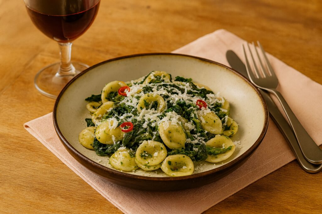 Orecchiette mit Cime di Rapa, scharfem Chili und geriebenem Käse, angerichtet auf einem hellen Holztisch mit Rotweinglas.