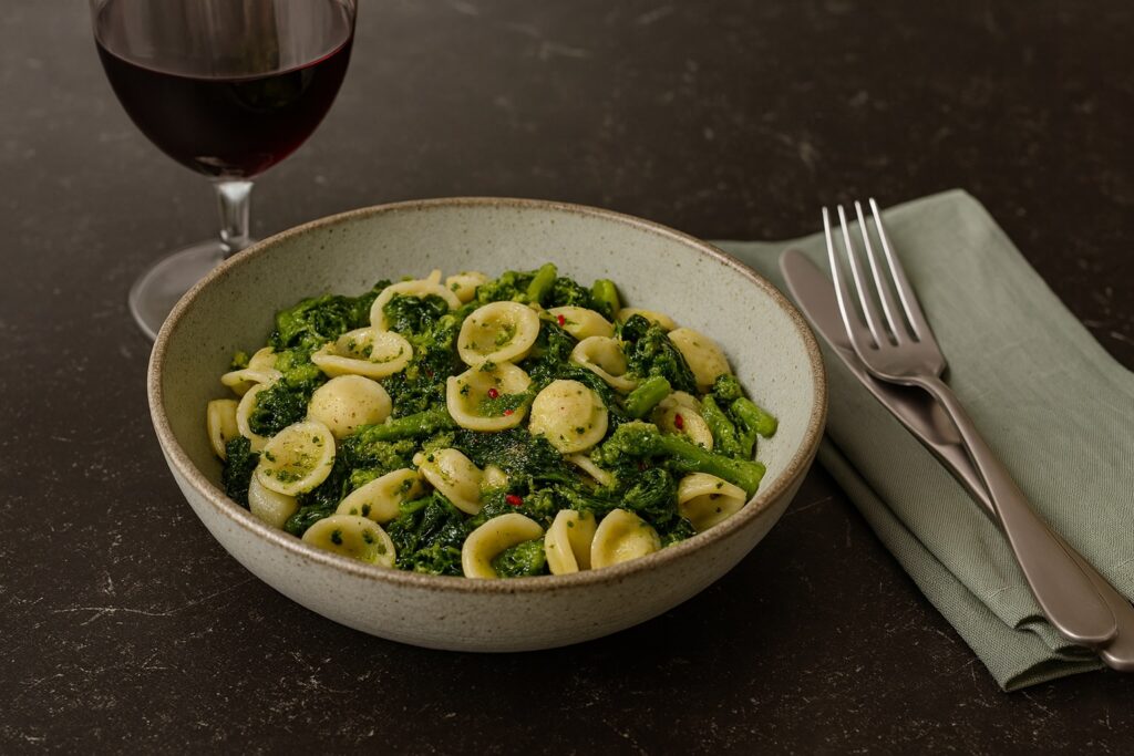 Italienisches Gericht mit Orecchiette und grünem Gemüse, serviert mit Rotwein auf dunklem Steinuntergrund.