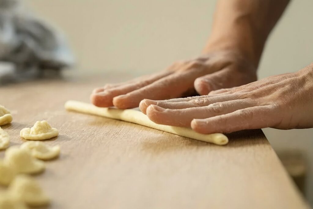 Zwei Hände rollen frischen Nudelteig auf einem Holztisch für die Herstellung von Orecchiette.