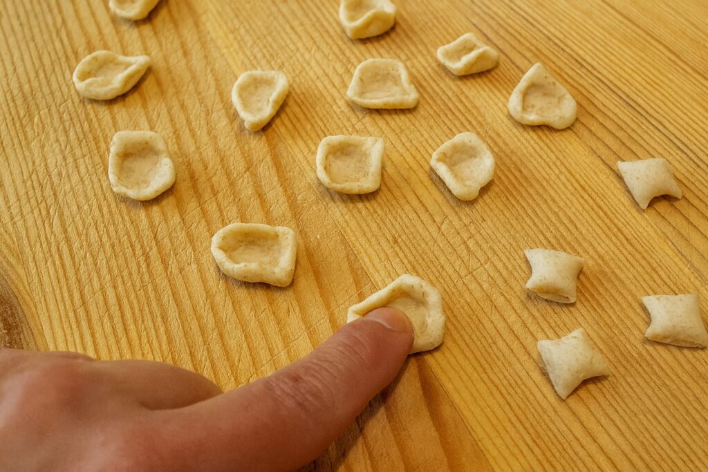 Finger drücken kleine Teigstücke zu Orecchiette-Formen auf einem Holzbrett.