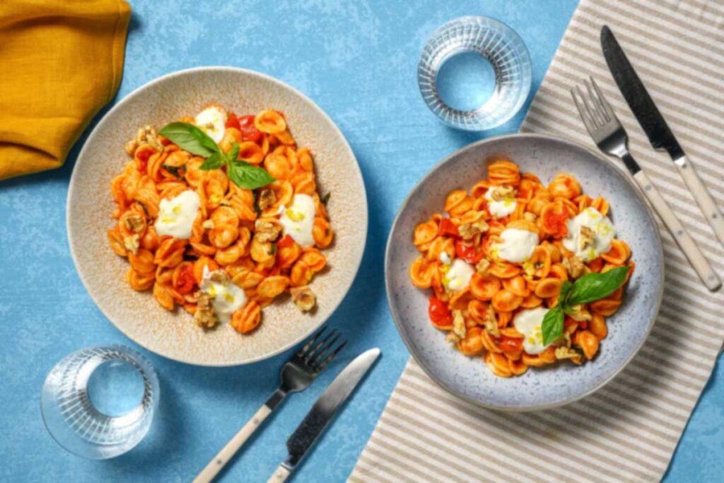 Zwei bunte Teller mit Orecchiette in Tomatensauce, garniert mit Mozzarella, Walnüssen und Basilikum, auf einem blauen Untergrund angerichtet.