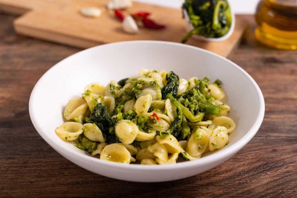 Orecchiette mit Cime di Rapa (Stängelkohl) in einer weißen Schüssel auf einem Holztisch, im Hintergrund Knoblauch, Chili und Olivenöl.