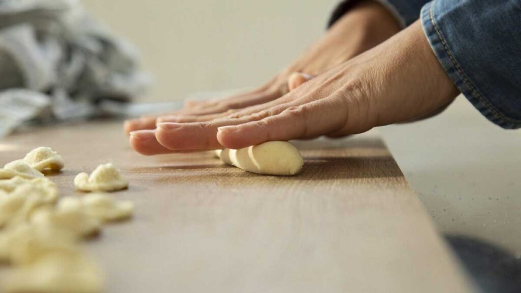 Nahaufnahme von Händen, die frischen Orecchiette-Teig auf einem Holzbrett rollen.