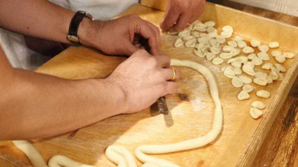 Zwei Personen bei der gemeinsamen Zubereitung von Orecchiette: Teig wird gerollt und mit dem Messer geschnitten.