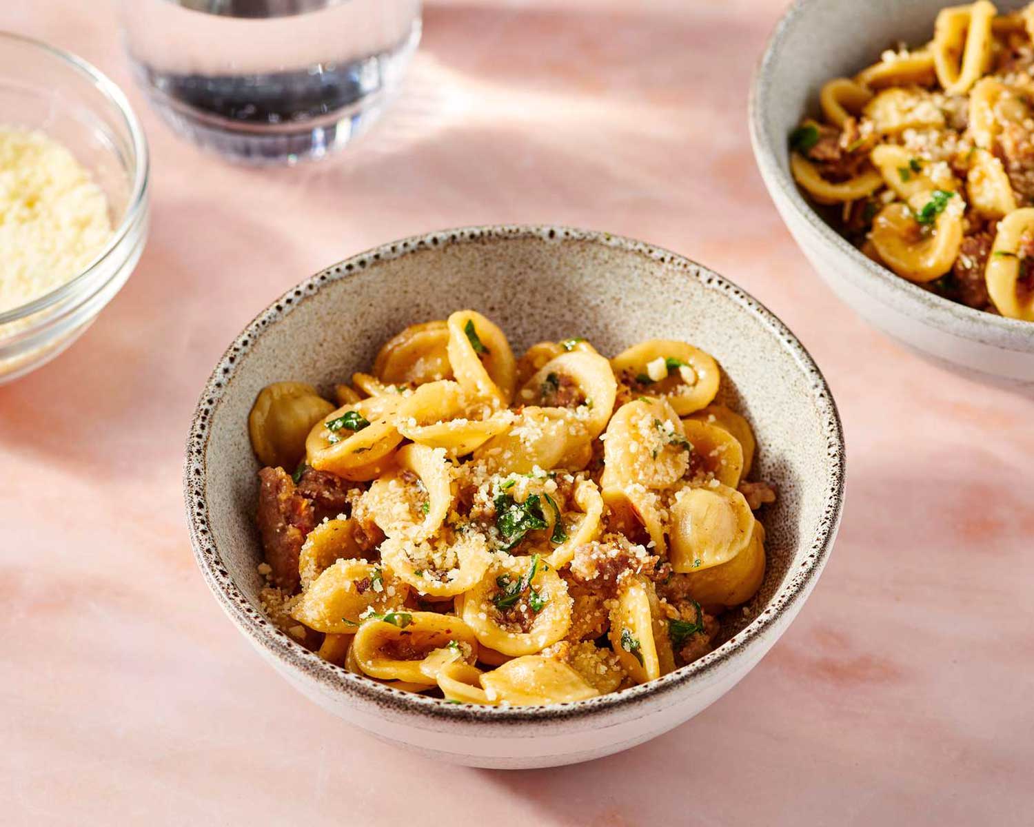 chale mit Orecchiette alla salsiccia – Pasta mit italienischer Wurst, Kräutern und geriebenem Käse auf rosa Untergrund.