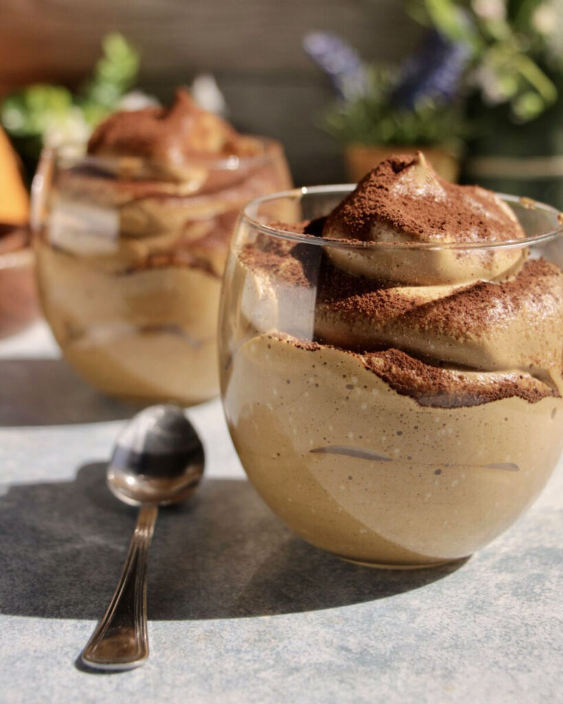 Luftig geschlagener Kaffee-Mousse in bauchigen Gläsern, serviert mit Löffel und Sonnenlicht – ein perfekter Genussmoment an warmen Tagen.