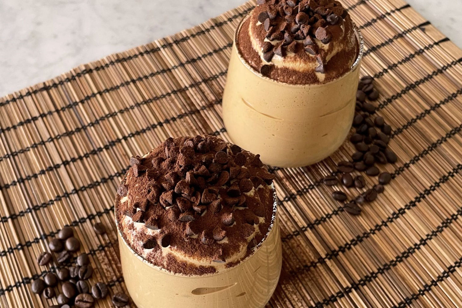 Zwei Gläser Spumone al Caffè mit Sahne, Kakaopulver und Schokosplittern garniert, serviert auf Bambusmatte mit Kaffeebohnen – Dessert mit Wow-Effekt.
