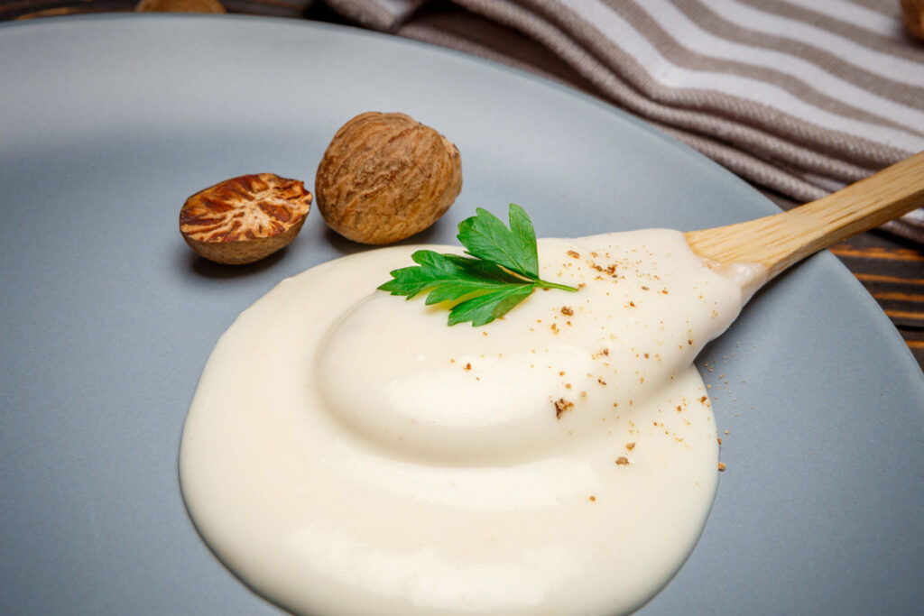 Nahaufnahme einer cremigen Besciamella-Sauce auf einem Teller, garniert mit Petersilie und frischer Muskatnuss