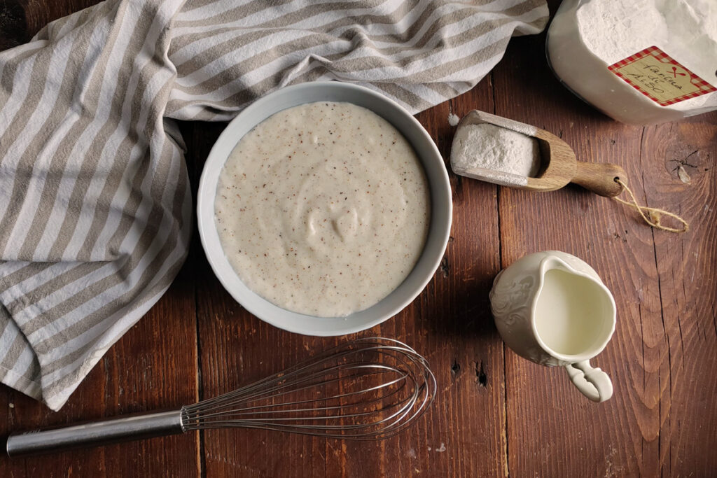 Schüssel mit frisch zubereiteter Béchamelsoße auf einem Holztisch, umgeben von Mehl, Milch und Muskatnuss – typische Zutaten der klassischen Sauce