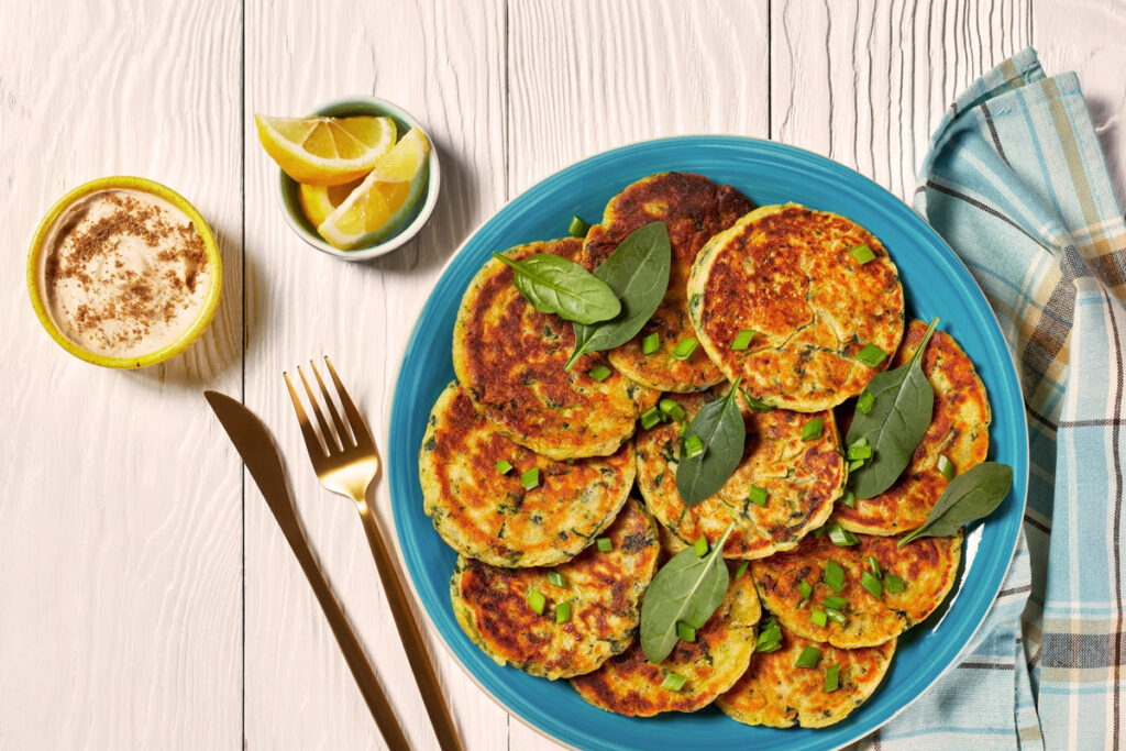 Frittelle di Ceci angerichtet auf einem blauen Teller, dekoriert mit Spinatblättern und Frühlingszwiebeln, serviert mit einer Schale Gewürzjoghurt und Zitronenspalten.